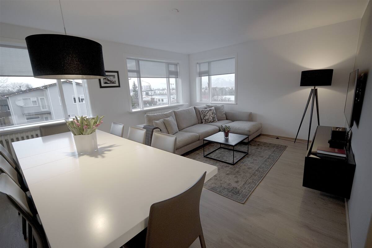 Uma sala de estar moderna com um sofá branco, uma mesa de jantar e uma mesa de centro. Há grandes janelas com vista para edifícios e uma montanha.