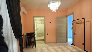 Une chambre avec un lit, un bureau et une chaise. On y trouve une porte qui donne sur une autre pièce, un radiateur et un portant à vêtements.