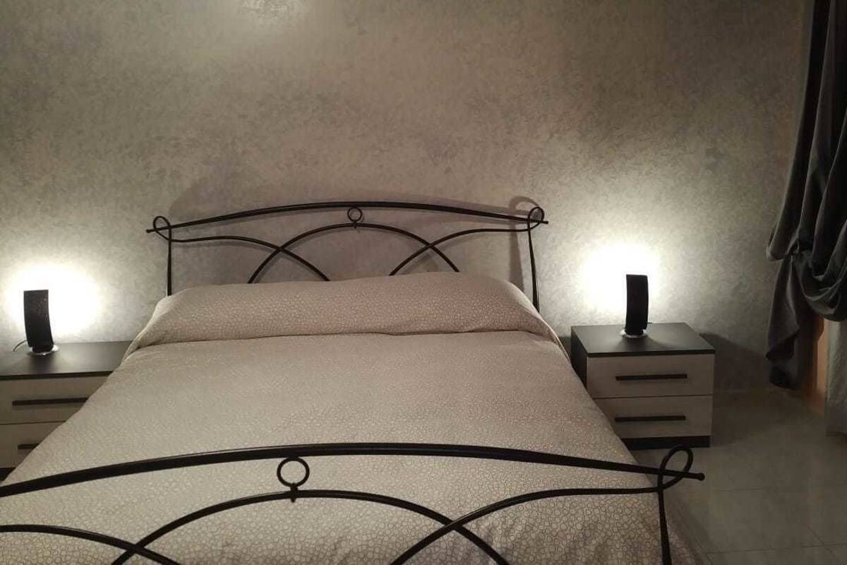 Ein Bett mit Metallgestell, zwei Nachttische mit Lampen und ein Vorhang rechts.