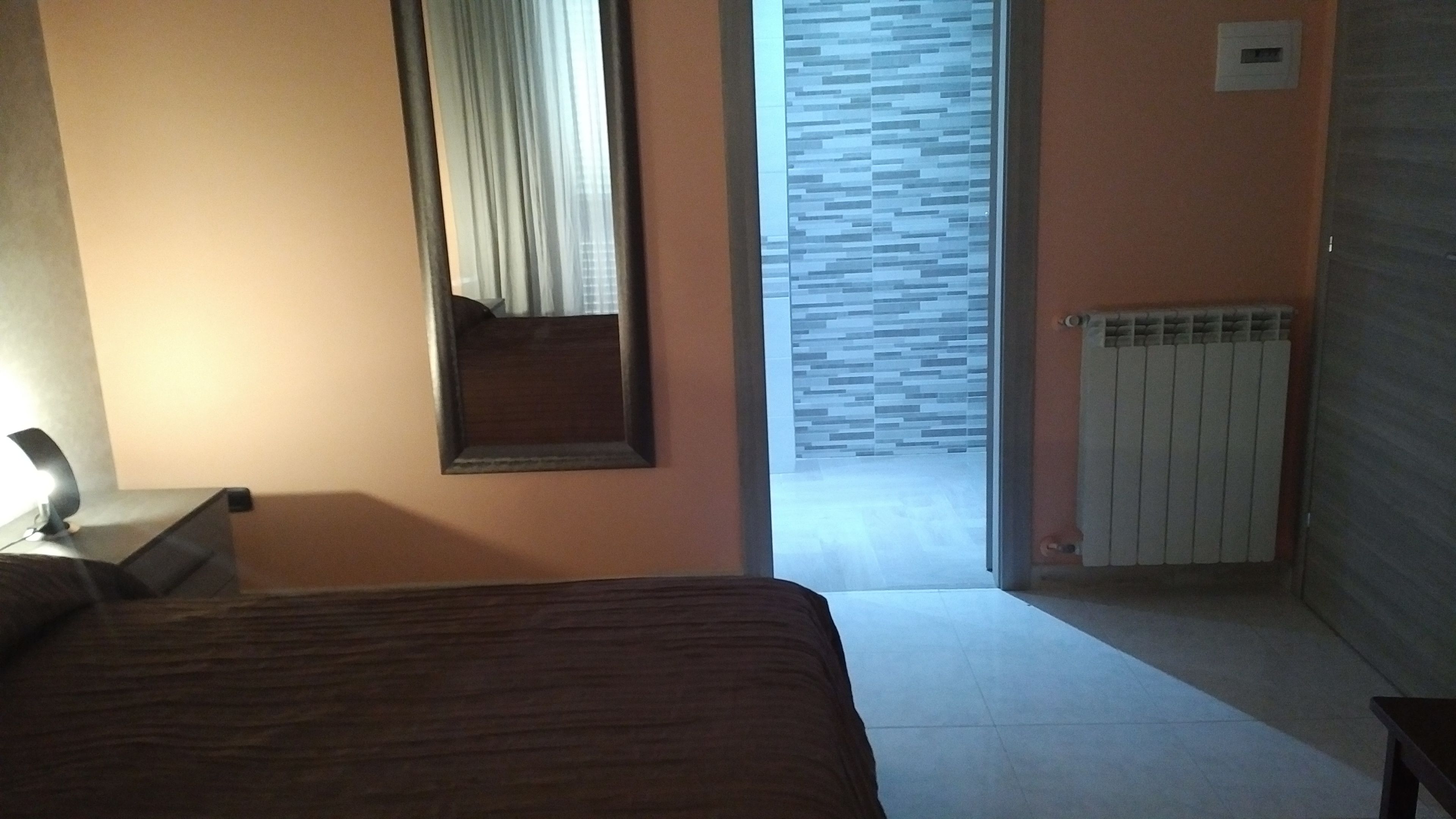 Een slaapkamer met een bed, een nachtkastje met een lamp, een spiegel, een radiator en uitzicht op een betegelde muur door een geopende deur.