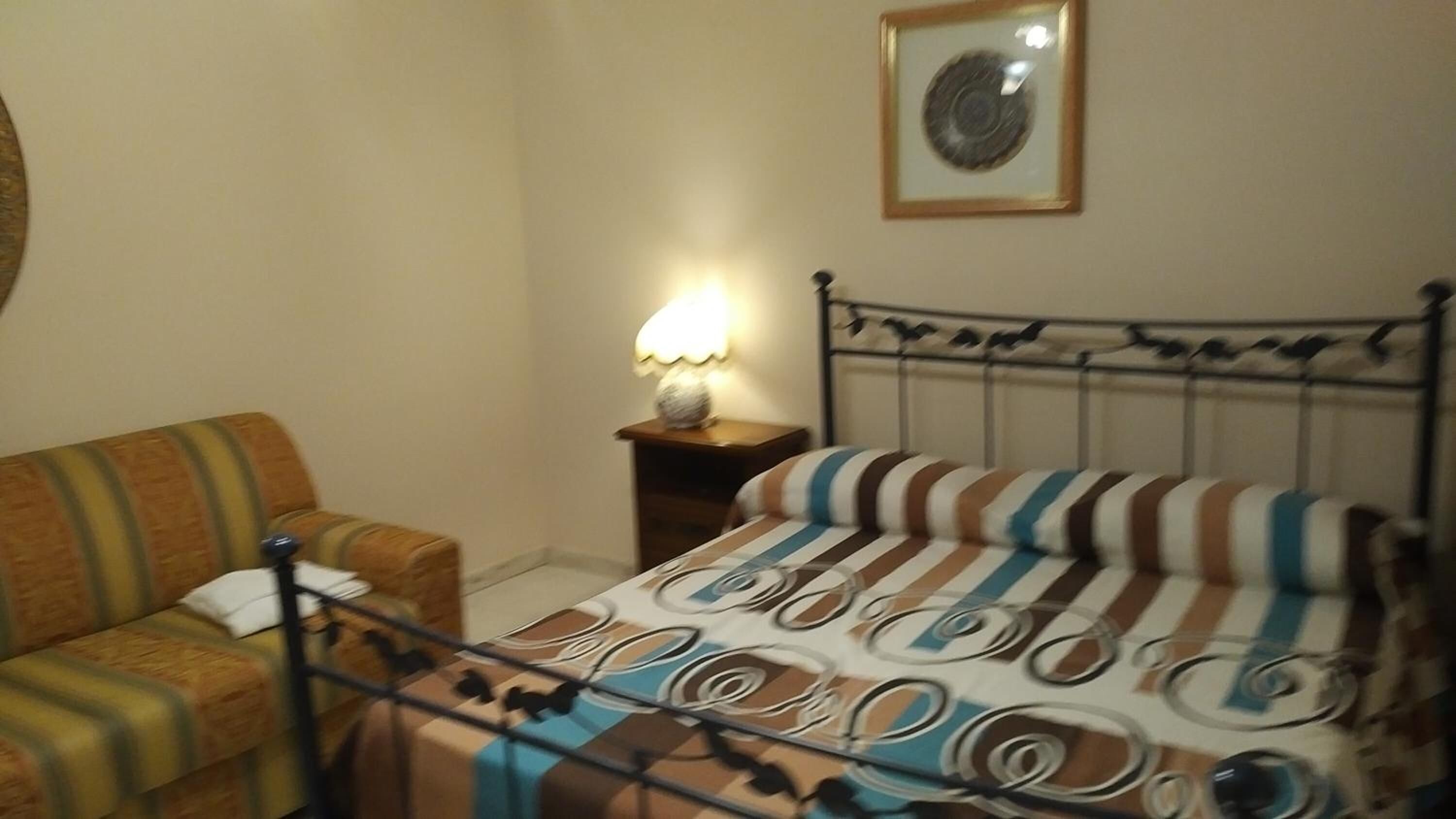A room with a bed, a chair, a nightstand with a lamp, and a framed picture on the wall.