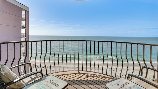 Un balcón con vistas a la playa y al océano. Hay dos sillas y una mesa con un mantel individual con motivos playeros.