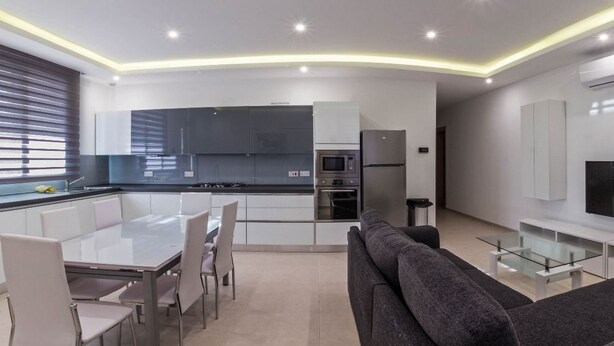 A modern kitchen with white cabinets, a dining table with chairs, a grey sofa, and a flat-screen TV on a white stand. - Ocean Gardens B3 (Naxxar)