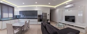 A modern kitchen with white cabinets, a dining table with chairs, a grey sofa, and a flat-screen TV on a white stand. - Ocean Gardens B3 (Naxxar)