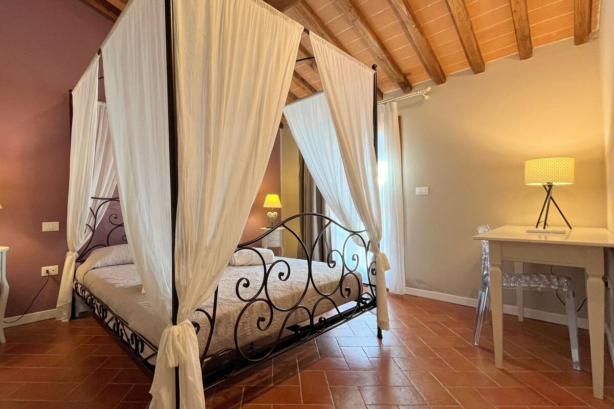 A bedroom with a four-poster canopy bed, two bedside tables with lamps, a small table with a lamp, and a fireplace.