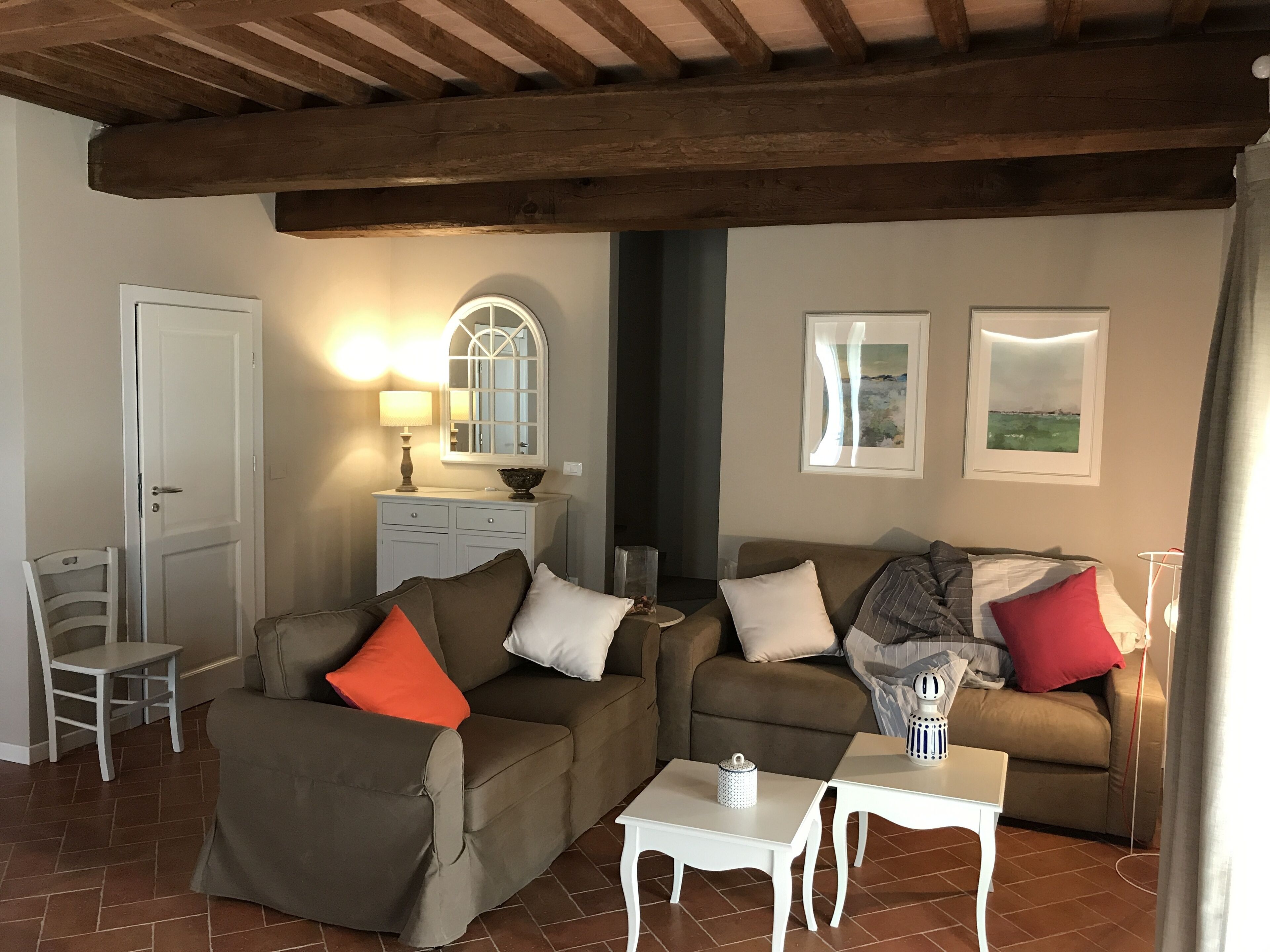 A cozy living room with a brown sofa, a white side table, and a white chair. There is a wooden ceiling and a white door.