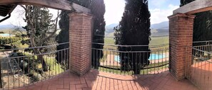 A balcony with a brick floor and metal railing, overlooking a pool and a landscaped garden with tall trees and a distant mountain range. - Borgo del Silenzio (Lajatico)