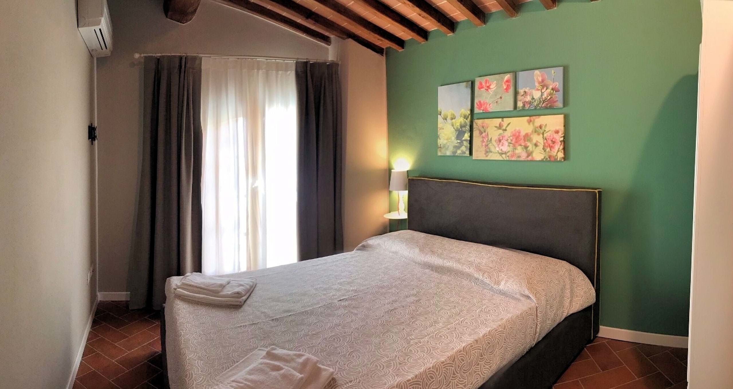 A bedroom with a bed, curtains, a wall-mounted lamp, and floral artwork.