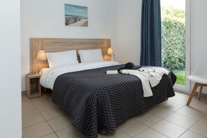 Uma cama bem arrumada com um edredom estampado, duas luminárias de cabeceira, uma pequena mesa de cabeceira e vista para o verde através de uma janela.