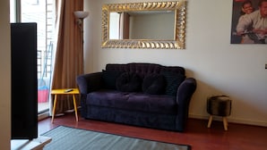 Ein Wohnzimmer mit einem dunkelvioletten Sofa, einem kleinen gelben Beistelltisch, einem großen Spiegel mit aufwendigem Goldrahmen, einer Zeitschrift an der Wand und einem Fernseher auf einem Lowboard.