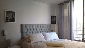 Ein ordentlich bezogenes Bett mit einem gemusterten Überwurf, einem gepolsterten Kopfteil und zwei Kissen. Ein kleiner Nachttisch mit Lampe und Telefon steht daneben. Über dem Bett hängen zwei gerahmte Bilder.