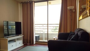 Ein Wohnzimmer mit einem Flachbildfernseher auf einem weißen Standfuß, einem dunklen Sofa und einer Glastür, die auf einen Balkon führt.