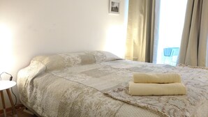 Ein ordentlich bezogenes Bett mit einer gemusterten Tagesdecke und einem gefalteten Handtuch, ein kleiner Nachttisch mit einer Lampe und ein Fenster mit Vorhängen.