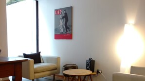 Ein Wohnzimmer mit einer Couch, einem runden Holztisch, einem kleinen Hocker, einem Poster an der Wand und einem Lautsprecher.