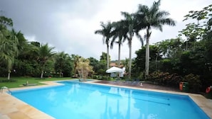 A swimming pool surrounded by palm trees and greenery, with a white umbrella and lounge chairs nearby.