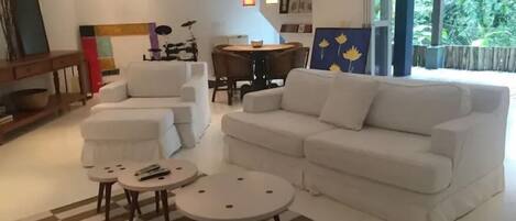 Un salon moderne avec un canapé blanc, deux fauteuils, une table basse et une table à manger. Un tableau orne le mur, une batterie est visible au fond, et une fenêtre offre une vue sur la verdure.