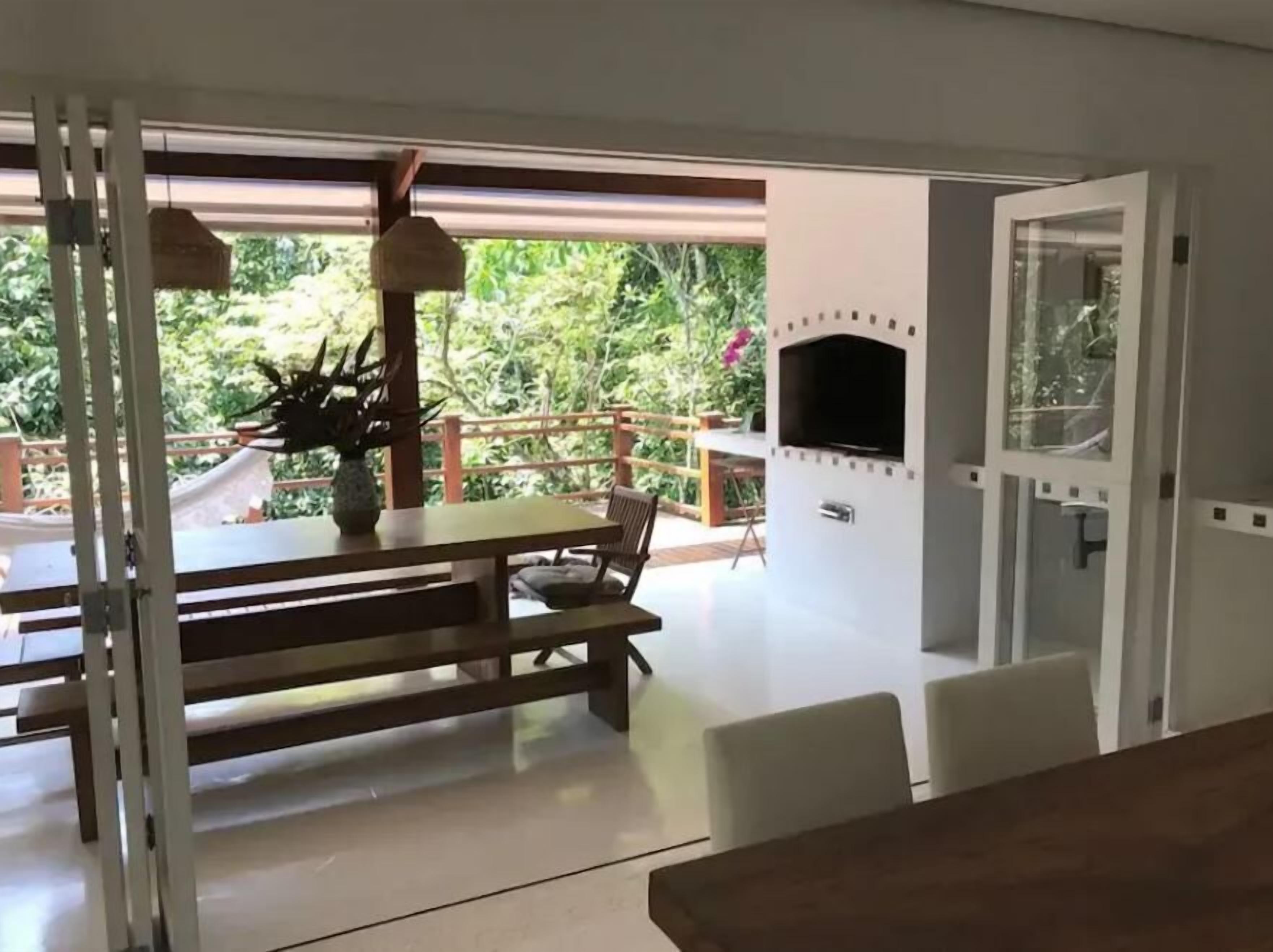 A dining area with a table and chairs, a TV mounted on the wall, and a view of greenery outside.