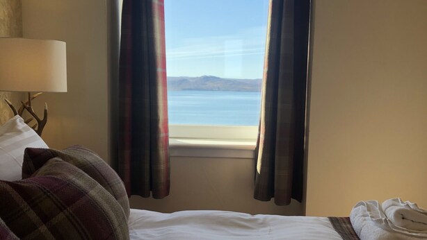 A hotel room with a bed, a lamp, a window with curtains, and a view of the sea. - The Millcroft Hotel (Gairloch)
