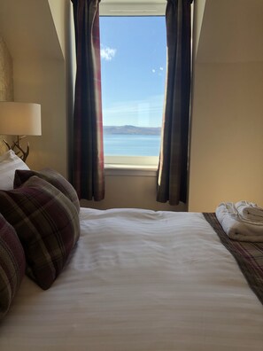 A hotel room with a bed, a lamp, a window with curtains, and a view of the sea. - The Millcroft Hotel (Gairloch)