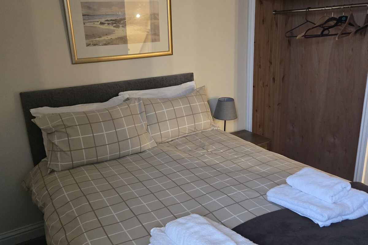 Een net opgemaakt bed met een geruit beddekleed, twee kussens en twee opgevouwen handdoeken op het bed.