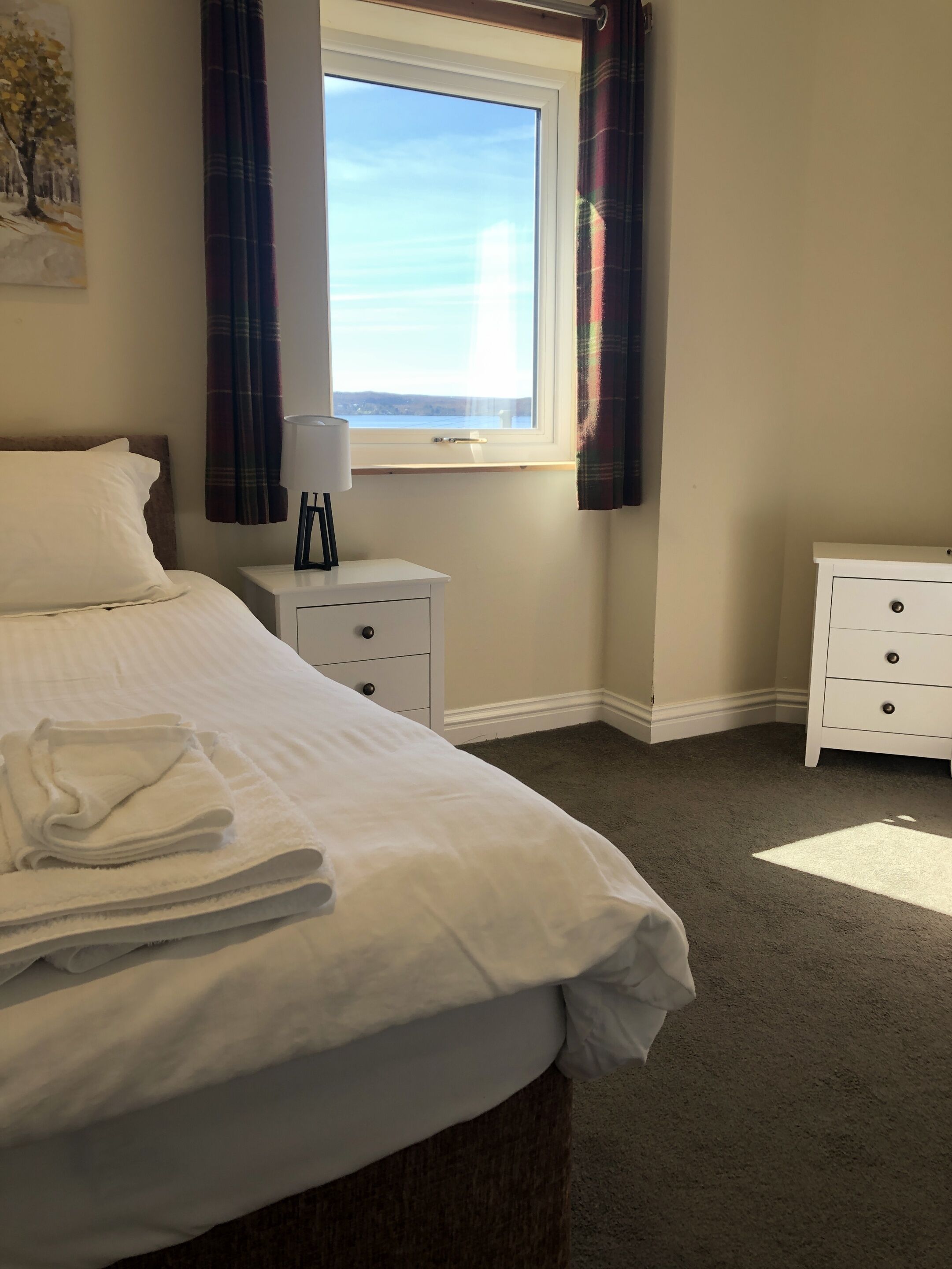 Habitación de hotel con cama, mesita de noche, lámpara, ventana con cortinas y vista al mar.