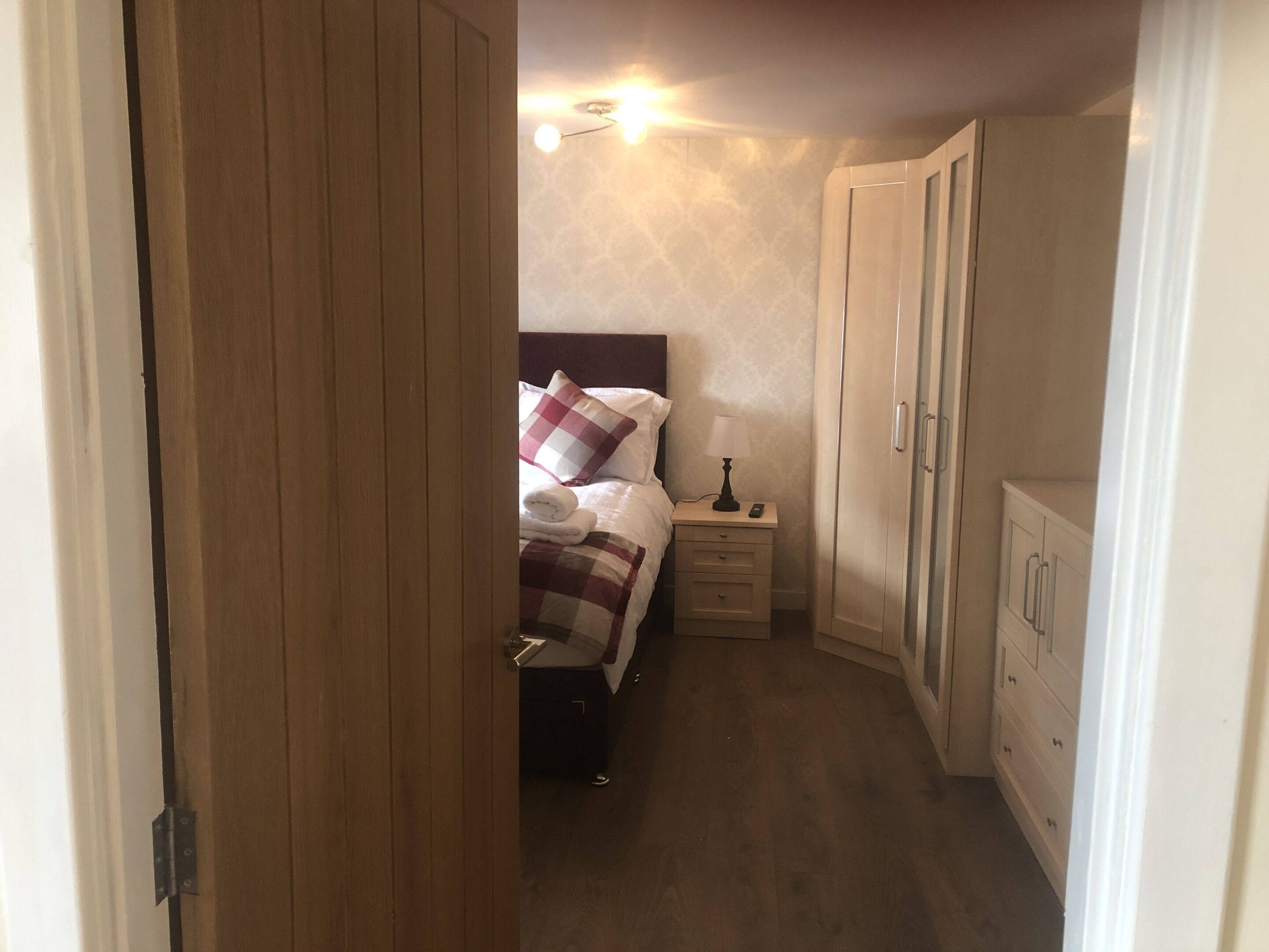 A narrow hotel room with a bed, bedside table, lamp, and wardrobe.