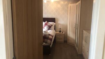 A narrow hotel room with a bed, bedside table, lamp, and wardrobe.