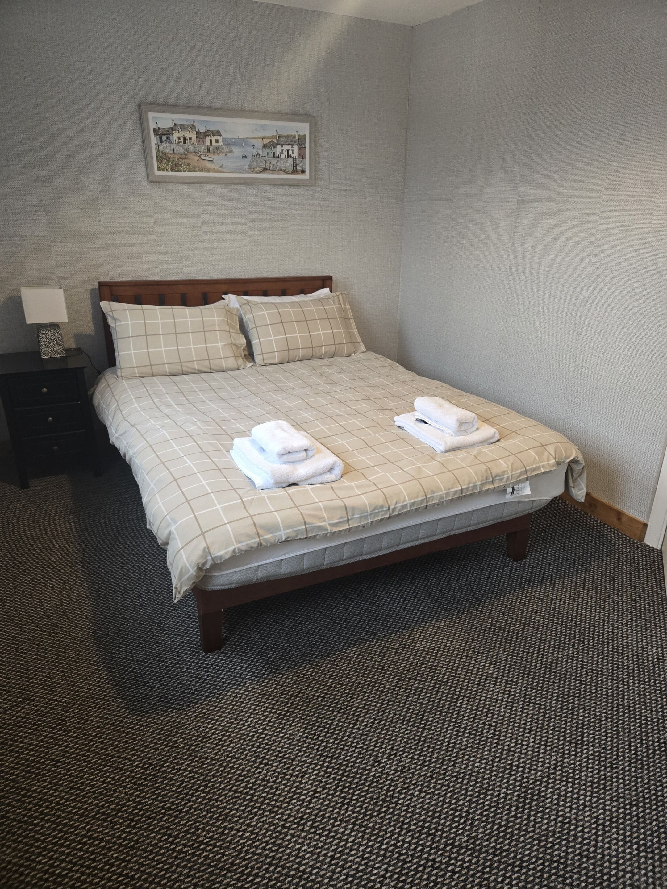 A neatly made bed with a checkered bedspread, two pillows, and two folded towels. There is a framed picture on the wall above the bed.