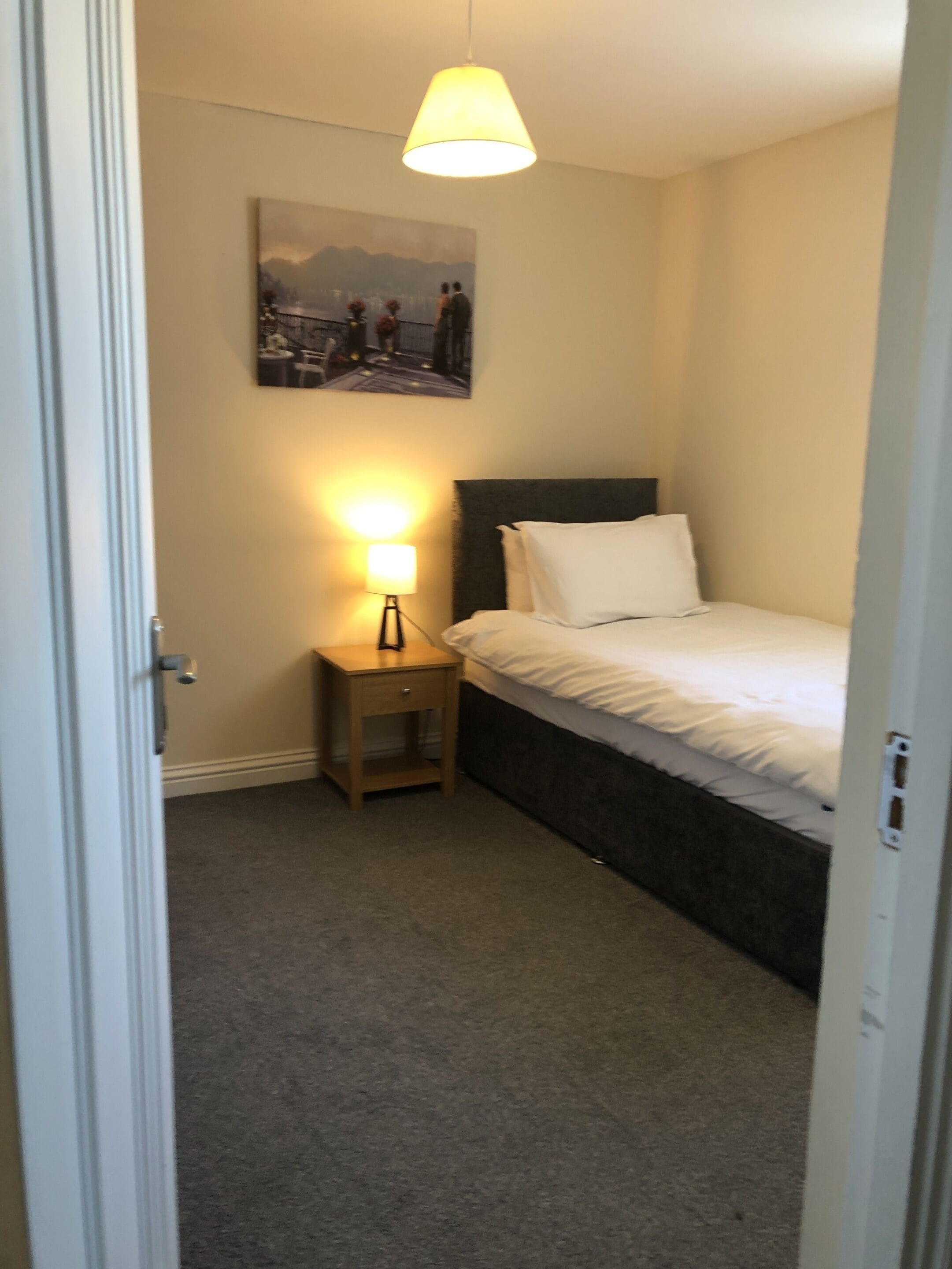 A small, neatly arranged bedroom with a single bed, a bedside lamp, and a wall-mounted picture.