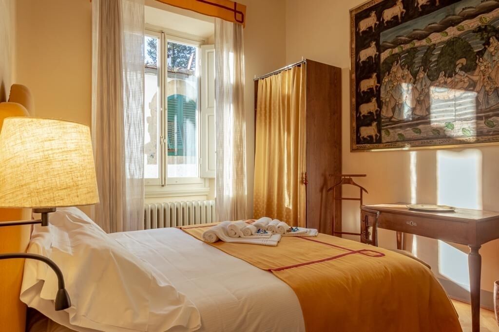 A hotel room with a bed, a window with curtains, a radiator, a table with a lamp, and a painting on the wall.