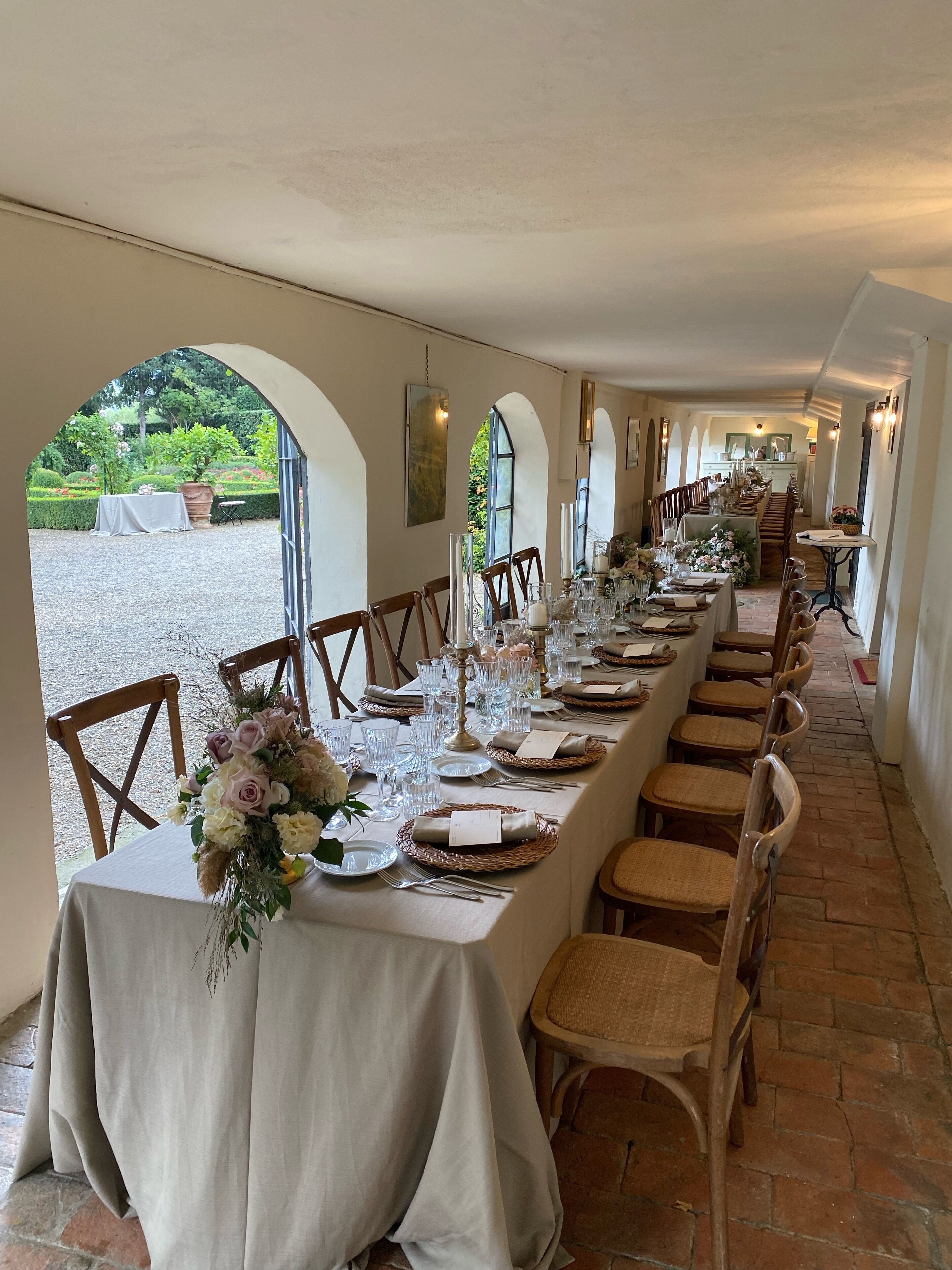 Ein langer Esstisch mit Stühlen, weißen Tischdecken und floralen Tischdekorationen in einem Gang mit Rundbogenfenstern und Blick auf Grünfläche.