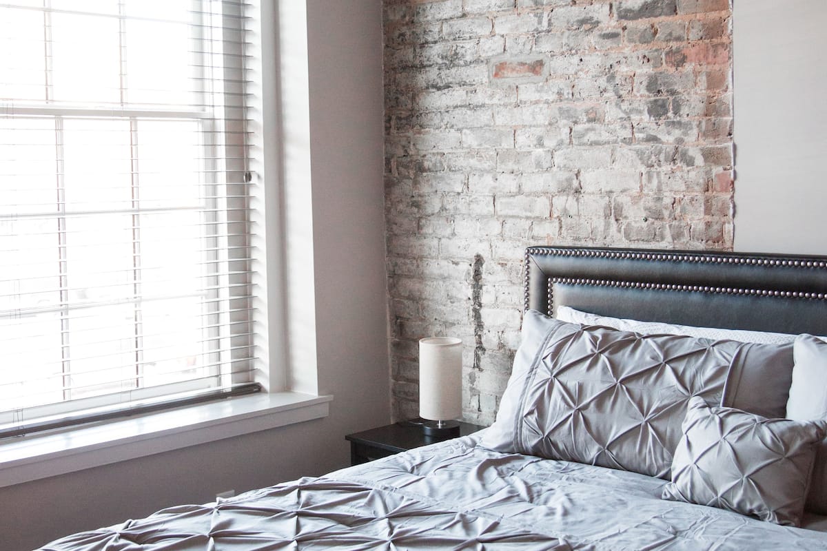 A bedroom with a brick wall, a large window with blinds, a bed with a patterned bedspread, and a bedside lamp.