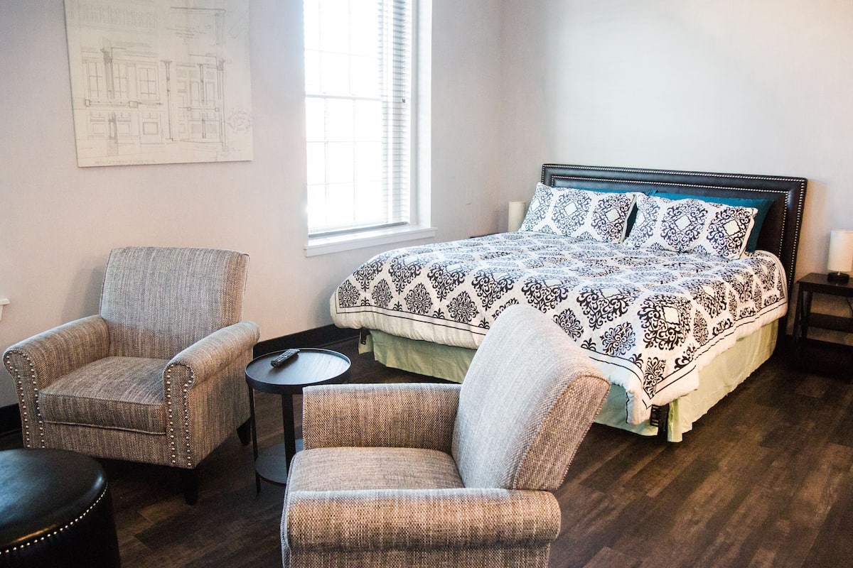 A bedroom with a bed, two armchairs, a small table, and a window with blinds.