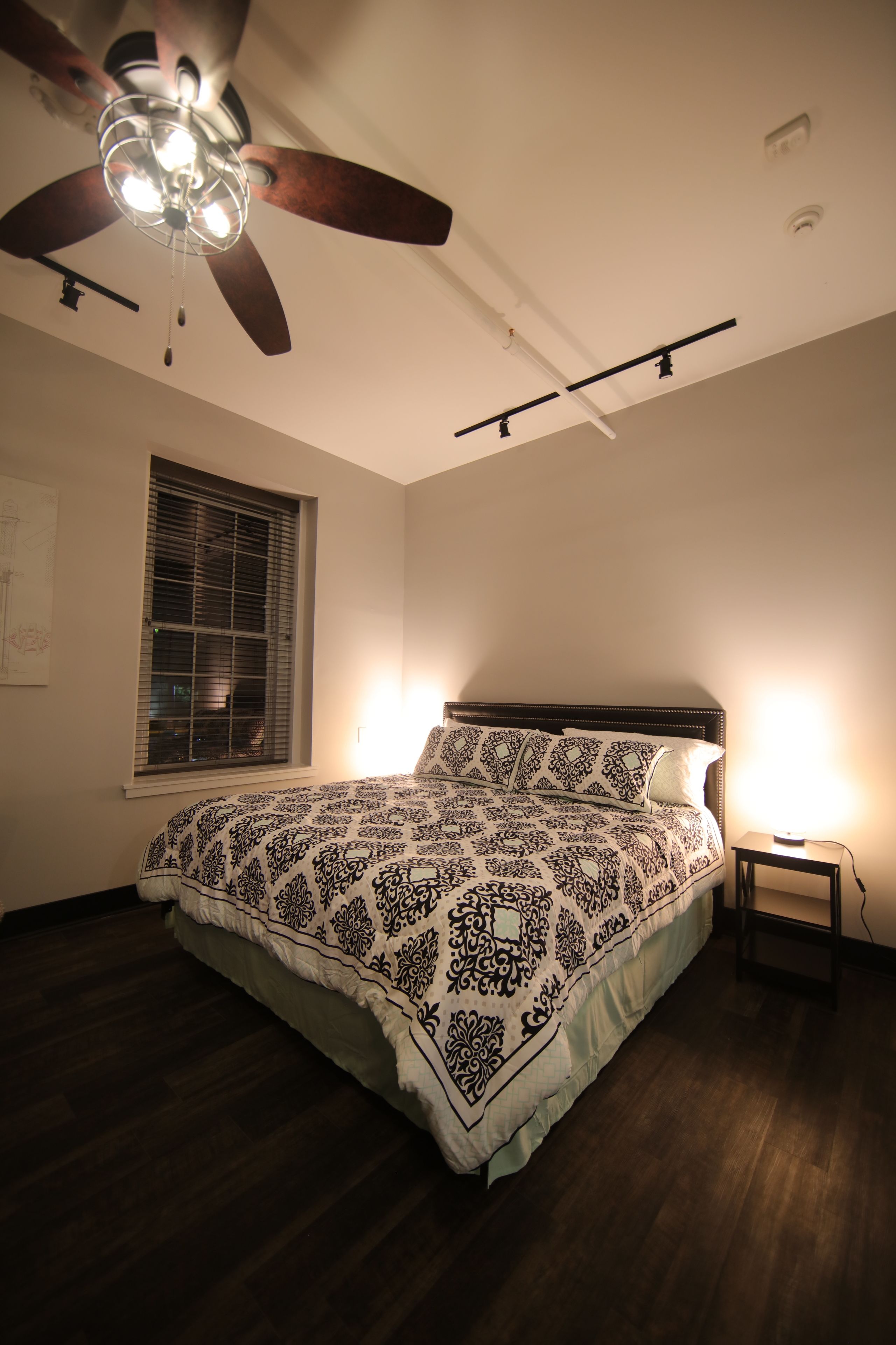 A bedroom with a bed, ceiling fan, window with blinds, and bedside lamp.