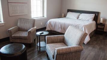 A hotel room with a large bed, two armchairs, a small round table, and a black ottoman.