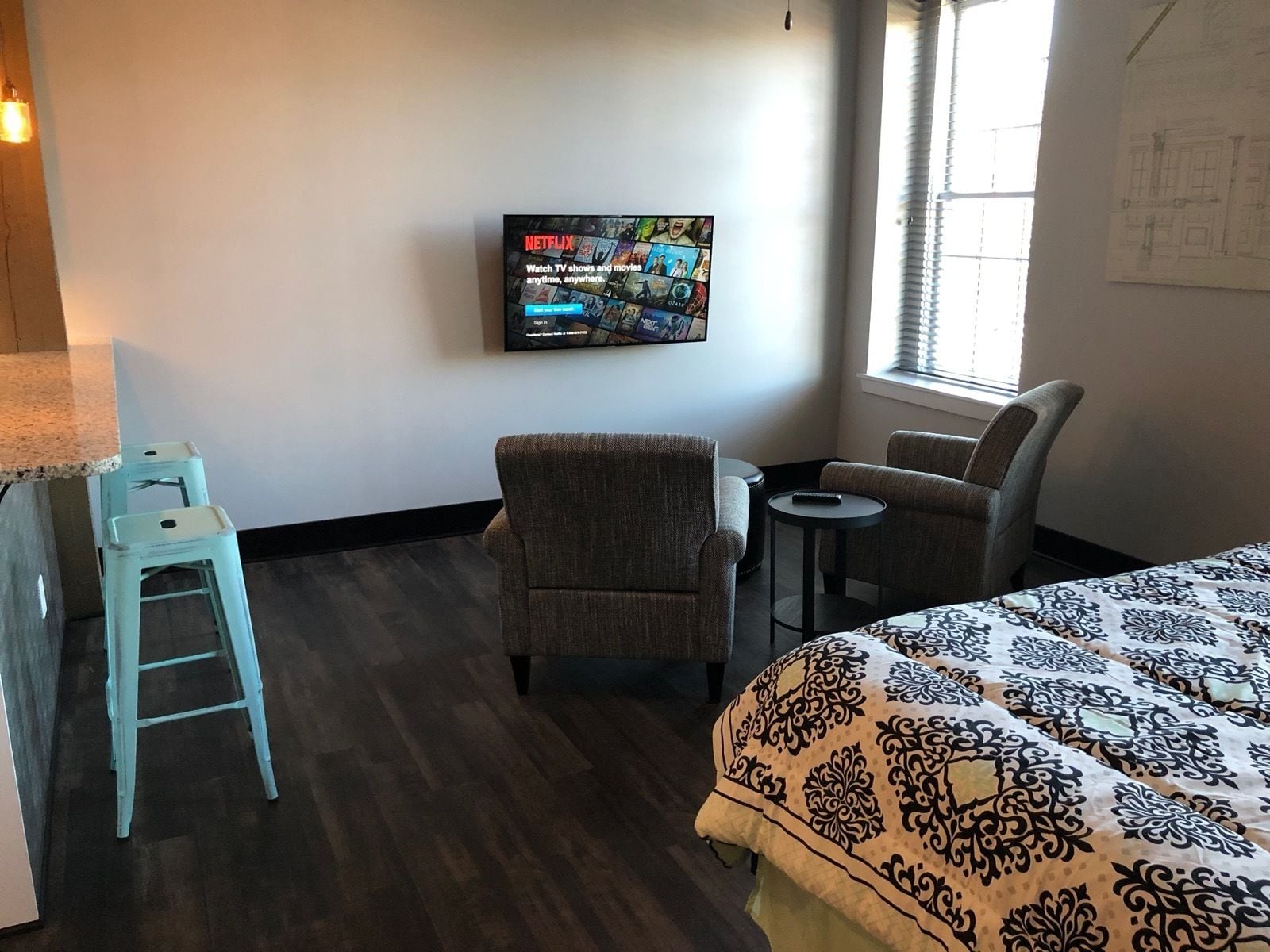 A hotel room with a bed, two armchairs, a small table, a TV mounted on the wall, and a window with blinds.