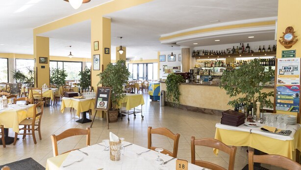 Un restaurante con paredes amarillas, varias mesas con manteles blancos y una zona de bar con diferentes botellas.