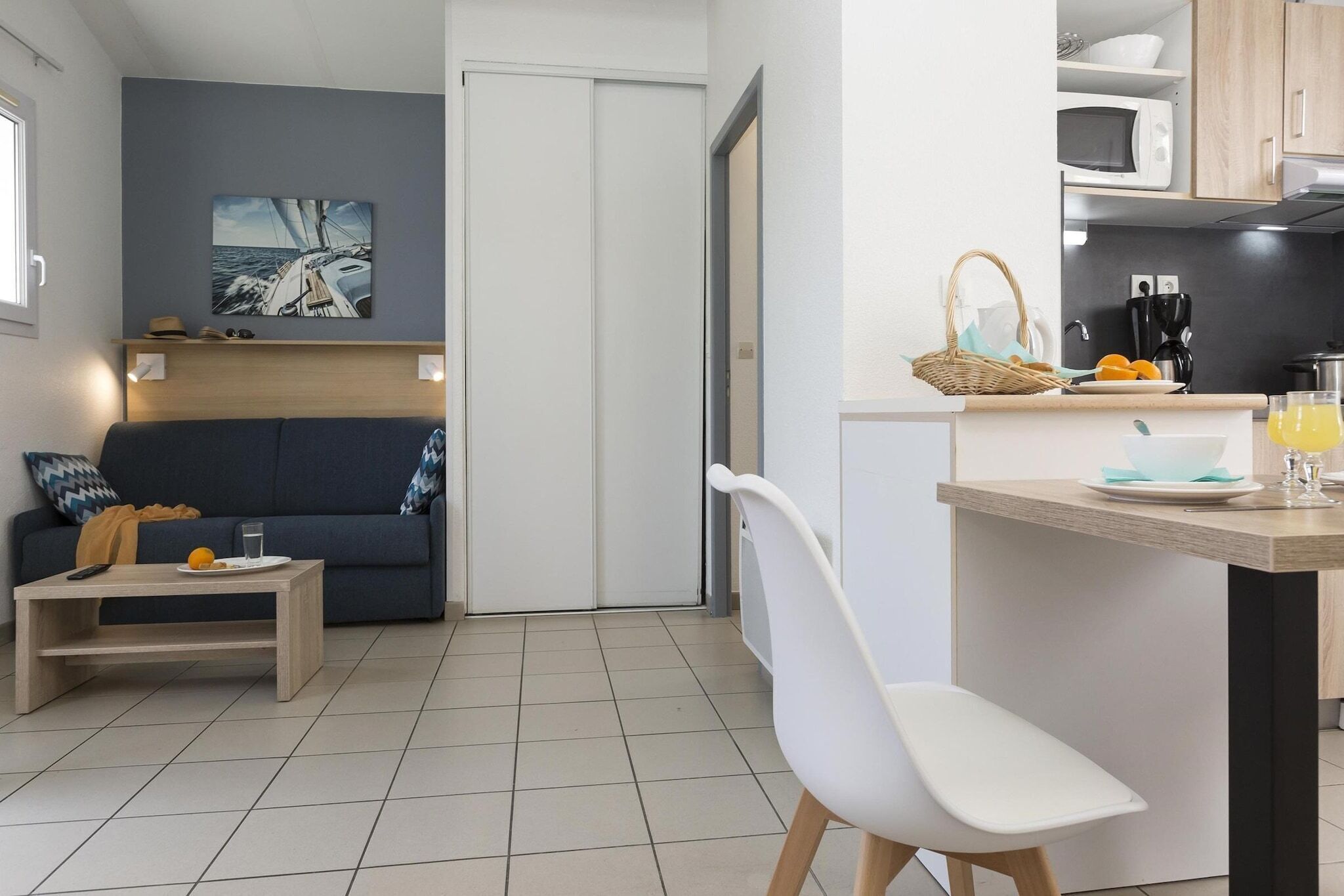 Un espace de vie compact comprenant un canapé, une petite table, une chaise blanche, un coin cuisine avec un four à micro-ondes, et une table à manger avec un panier et un verre de jus d’orange.