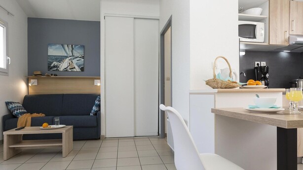 Espaço compacto com sofá, uma mesinha, uma cadeira branca, área de cozinha com micro-ondas e mesa de jantar com cesta e um copo de sumo de laranja.