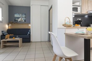 Un espace de vie compact comprenant un canapé, une petite table, une chaise blanche, un coin cuisine avec un four à micro-ondes, et une table à manger avec un panier et un verre de jus d’orange.
