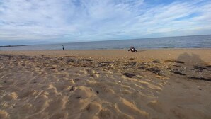 Uma praia com céu claro, uma pessoa caminhando e outra sentada na areia.