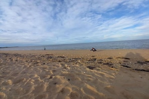 Ein Strand mit blauem Himmel, eine Person geht spazieren und eine andere sitzt im Sand.