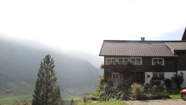 A two-story house with a tiled roof, surrounded by greenery and a foggy valley. - Vacation apartment Helma - Holiday apartment 1 (Blons)