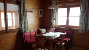 A wooden cabin interior with a dining table, chairs, a bench, and two windows with curtains. - Vacation apartment Helma - Holiday apartment 1 (Blons)