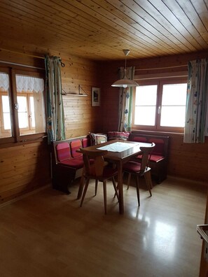 Ein Holzblockhaus-Interieur mit Esstisch, Stühlen, einer Bank und zwei Fenstern mit Vorhängen.