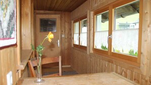 A wooden cabin interior with a wooden bench, a table, a vase with a single yellow flower, and windows with lace curtains. - Vacation apartment Helma - Holiday apartment 1 (Blons)