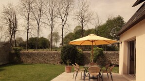 Une terrasse avec une table et des chaises sous un parasol jaune, entourée d’un mur de pierre et d’arbres.