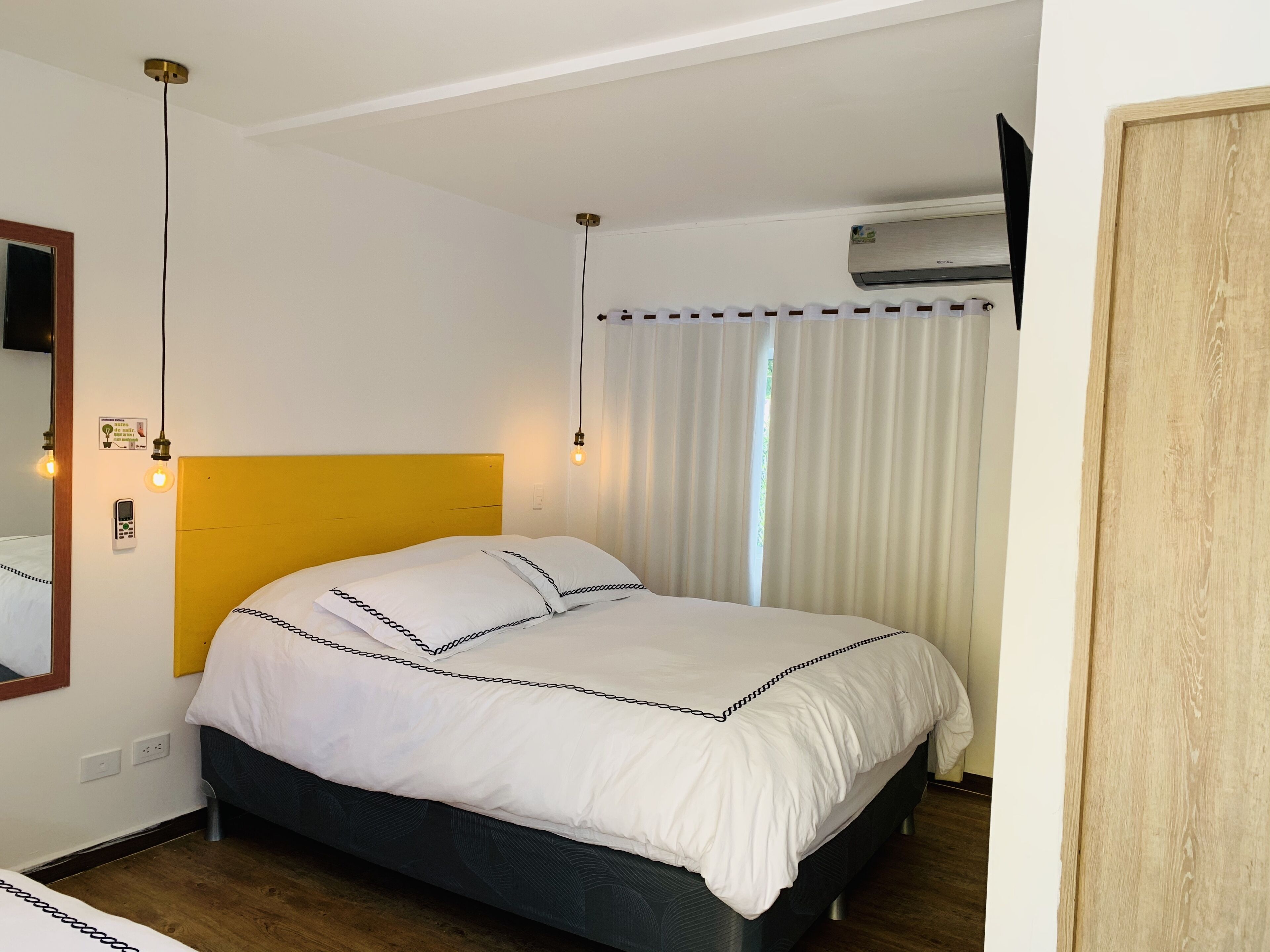 Hotellihuone, jossa on sänky, yöpöytä, peili, seinälle asennettu ilmastointilaite ja ikkuna, jossa on verhot.