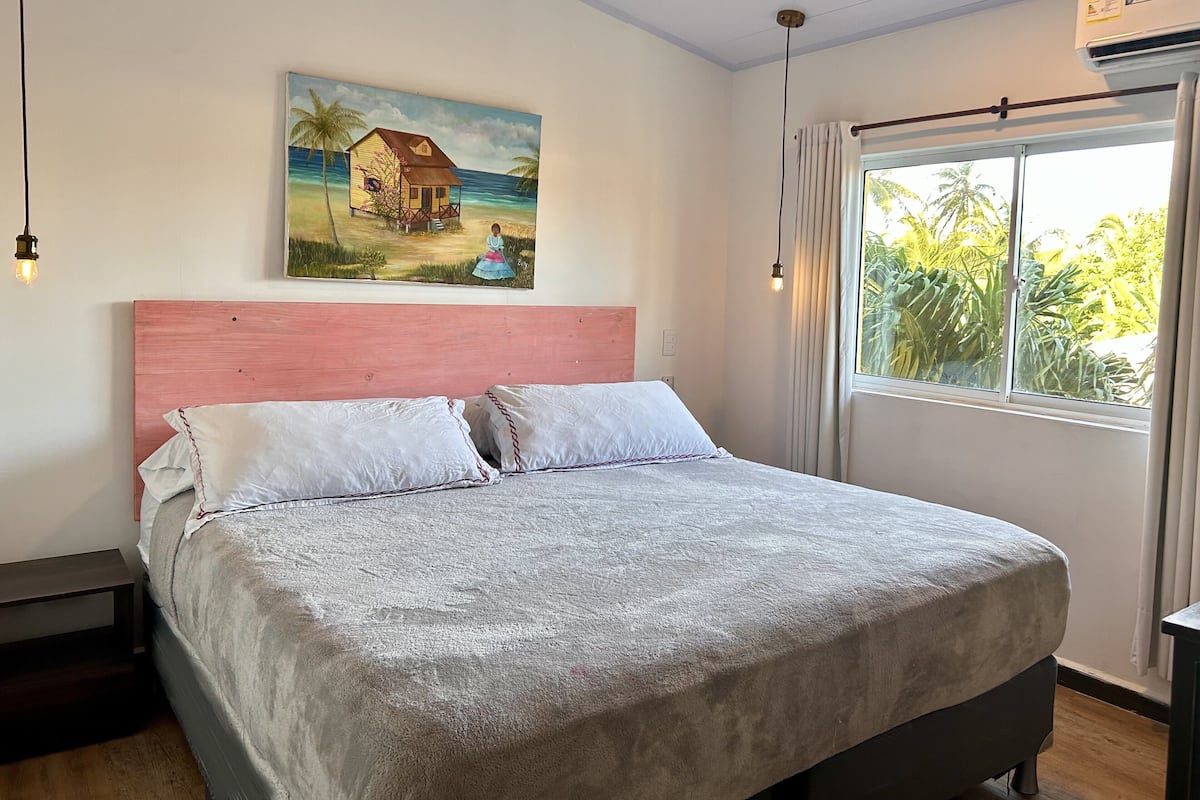 A bedroom with a bed, a painting of a house by the sea, a window with curtains, and a ceiling light.