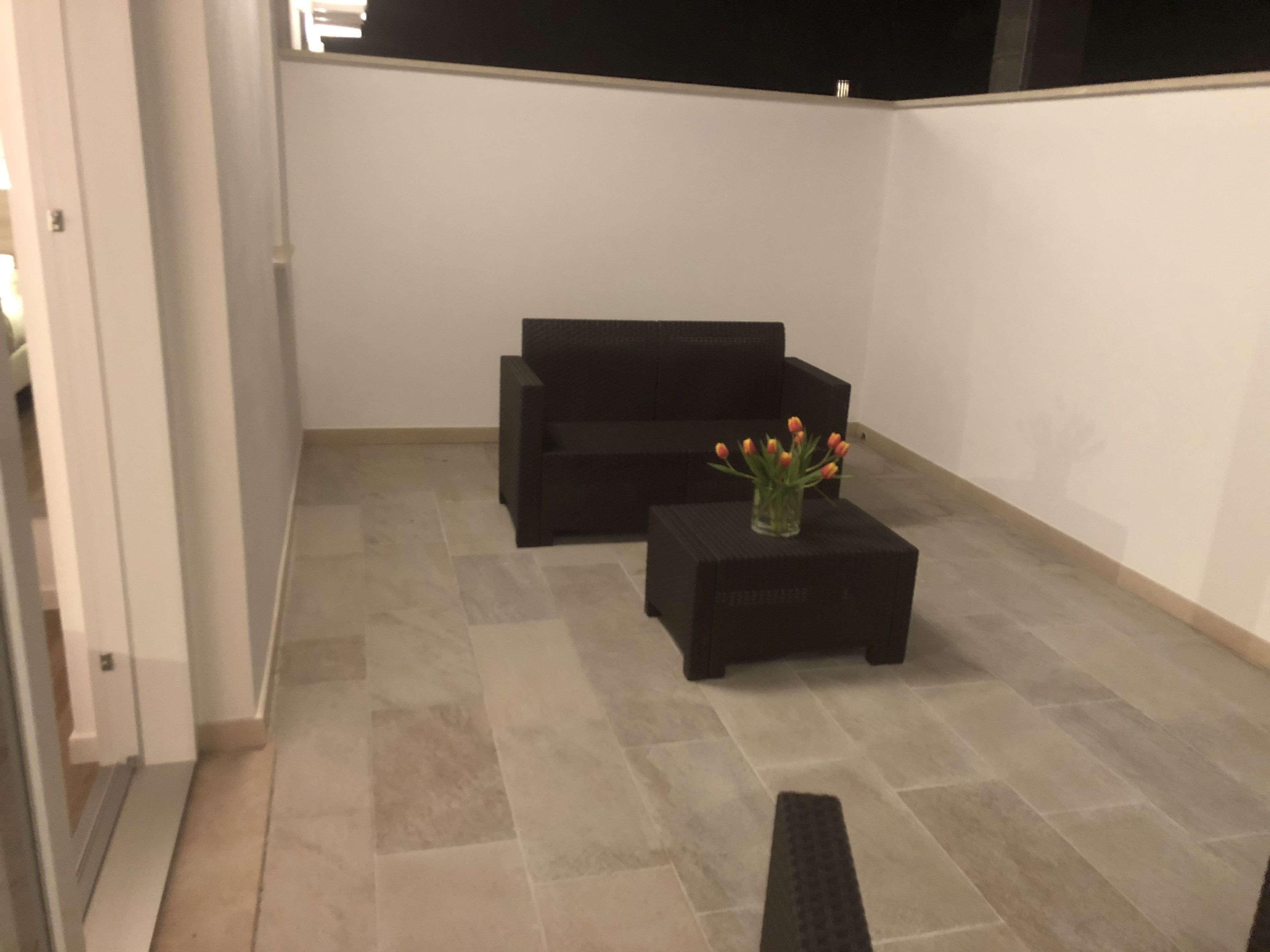 A small, enclosed space with a tiled floor, a black sofa, a small table with a vase of flowers, and a door on the left.